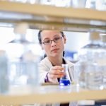 Female lab researcher reaches for a beaker in a science lab – photographed for a biotech annual report